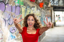 Cheerful Woman Gesturing Peace Sign Near Graffiti Wall On Footpath