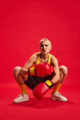 Cool sporty and slim woman in boxing gloves and sport cloths sitting emotionally looking at camera against red studio background. Concept of hobby, active-lifestyle, self expression.