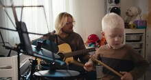 A Little Albino Boy With White Hair With A Short Haircut In A Brown Jacket Plays On An Electronic Drum Set Using Special Drum Sticks Together With His Father, A Blond Man In Glasses With A Beard Who