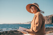 Serene retirement lifestyle. Smiling senior woman in outdoors enjoying retirement sitting face the sea looking at horizon over water reading a book