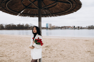 Wall Mural - Attractive brunette woman walking on the beach shore in moody cloudy windy weather with bouquet of red tulips flowers, dressed in white suit jacket. International Women`s Day 8th March concept