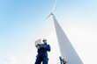 © narong - Professional technician man stand with hold laptop and stand near base of windmill or wind turbine with his co-worker in the back.