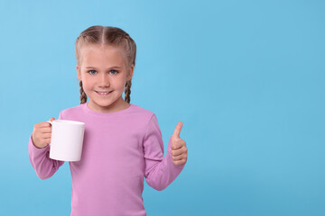 Sticker - Happy girl with white ceramic mug showing thumbs up on light blue background, space for text