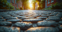 Close-up View Of A Cobblestone Street Leading Towards A Glowing Sunset Between Buildings With Trees And Warm Light