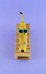 Wall Mural - Banana with sunglasses resting on the wooden beach bed. Summer vacation concept on purple background