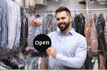 Wall Mural - Dry-cleaning service. Happy worker holding Open sign at counter indoors