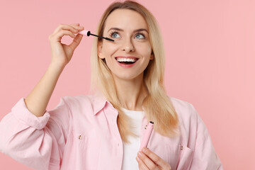 Wall Mural - Beautiful woman applying mascara on pink background