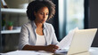 © Studio Nova - Focused woman with curly hair, wearing a white shirt, reading a document in a bright, home office setting, with a laptop open in front of her.