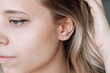 © Марина Демешко - Ear piercing. Cropped shot of a young blonde woman wearing four stud earrings on the earlobe. Jewelry with gemstones, accessories, a cross with fianits