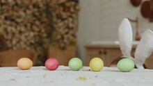 Multi Colored Painted Easter Eggs Lie On Table In House. Unrecognizable Child Is Hiding Behind Table And Playfully Swinging Headband With Bunny Ears. Playful Mood Of Boy In Costume On Easter Eve.