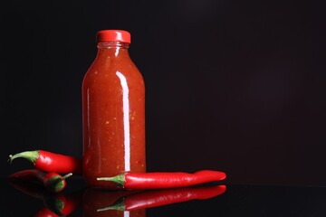 Wall Mural - Spicy chili sauce in bottle and peppers against dark background, space for text