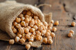 This photograph captures the rustic charm of chickpeas spilling from a burlap sack onto a weathered wooden surface, suggesting a harvest or market scene. 