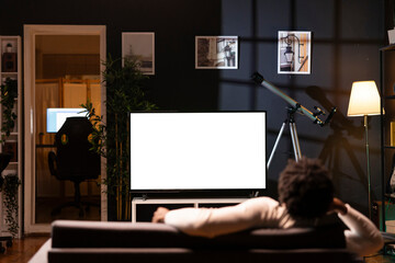 Homeowner bored at home, using isolated screen smart TV to binge series on subscription based streaming services. BIPOC man lounged on couch, enjoying VOD shows on mockup television display