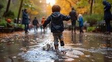Kid Walking In The Rain, A Little Boy Running Through A Puddle Of Water In A Park With Other People Watching Him From The Side