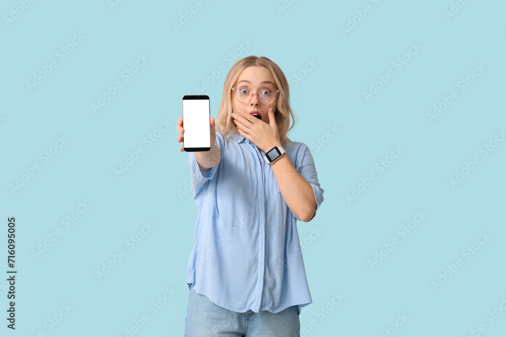 Shocked young woman showing smartphone with blank screen on blue background