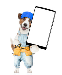 Poster - Funny Jack russell terrier puppy wearing blue cap and denim overalls with tool belt showing big smartphone with white blank screen in it paw. Isolated on white background
