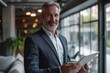 © sirisakboakaew - Happy middle aged business man ceo wearing suit standing in office using digital tablet.