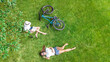© Iuliia Sokolovska - Young girls with bicycle in park, reading book and listening music with headphones, two student girls studying online outdoors sitting on grass near bike, aerial drone view from above