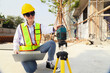 © ฺฺฺBoonterm - Female engineer foreman sitting holds laptop computer setup and checks  modern technology tools laser level and vertical marker (Plum Bob) on tripod at construction site, condominium building.