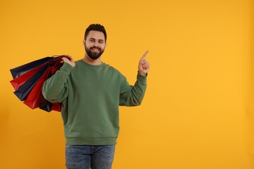 Sticker - Happy man with many paper shopping bags pointing at something on orange background. Space for text