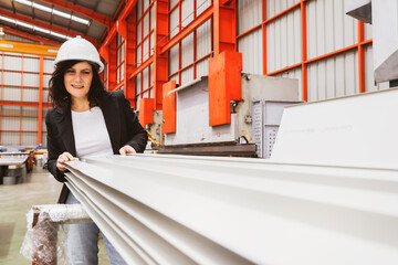 Female manager engineer wearing safety helmet inspects the production quality of white coated rolled metal roofing sheets, in a standardized and safe warehouse production facility.