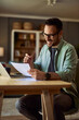 © Jelena - A happy adult male freelancer reading a document in his hand during an online meeting on a laptop.