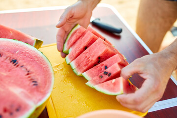 A person is cutting a ripe watermelon on a bright yellow cutting board, likely preparing a summery snack outdoors.