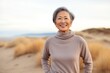 Portrait of a grinning asian woman in her 70s showing off a thermal merino wool top against a serene dune landscape background. AI Generation