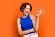 Photo of flirty pretty girl dressed blue top open mouth winking showing okey sign isolated orange color background