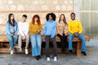 © JoseIMartin - A cheerful, multiethnic group of six friends enjoying a casual meet-up, seated on a wooden bench against a rustic brick wall backdrop
