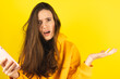 Mad and shock young brunette beautiful woman using mobile phone isolated on yellow background. Yelling unhappy woman hold mobile phone. Disappointed sad upset lady horrified impressed news. WTF. Oh no