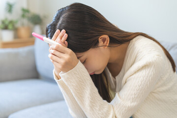 Sad, Disappointed motherhood asian young woman hand holding pregnancy test, upset ovulation problem, anxiety after result at positive sitting on couch at home. Unwanted Pregnant, contraception concept