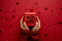 Close Up Female Hands Hold A Red Gift With A Red Ribbon On A Red Background Among Heart-shaped Confetti. Valentine's Day, Romance, Love, Anniversary Concept