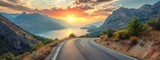 Fototapeta Natura - A winding road through a majestic mountain pass leads to a tranquil lake surrounded by towering trees, under a colorful sky at sunrise and sunset