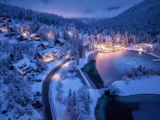 Wall Mural - Aerial view of fairy alpine village in snow, road, forest, Jasna lake, houses, street lights at winter night. Top view of mountains, illumination, snowy pine trees at twilight. Kranjska Gora, Slovenia