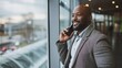 An African American businessman in an airport lounge, looking out the window with a smile while engaged in a conversation on his mobile phone, epitomizing modern business travel