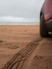 Side of a red car parked on a sandy beach with stunning view on the ocean. Travel and tourism concept. Renting vehicle for a trip theme. Cloudy sky and water surface in the background.