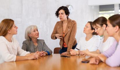 Canvas Print - Positive motivated young Hispanic woman team leader holding meeting with group of fellow female students of different ages sitting around desk, explaining details of new study project