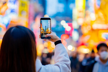 Asian female tourist Traveling and having fun. And she was taking photo with a cell phone camera at Night street with many restaurant around Tsutenkaku Tower in Shinsekai district of Osaka, Japan.