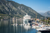 Fototapeta Tęcza - Low angle view of a large cruise ship docked at the port of Kotor in Montenegro, with the picturesque coastline, rocky peaks, red roofs of houses and a marina with boats.