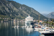 Low Angle View Of A Large Cruise Ship Docked At The Port Of Kotor In Montenegro, With The Picturesque Coastline, Rocky Peaks, Red Roofs Of Houses And A Marina With Boats.