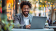 © SS Digital - A contented young Latino businessman sitting outside with a laptop. A happy Hispanic man studying or working in a city café, using a computer to do e-learning or hybrid work, and looking for a job onl