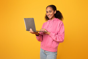 Wall Mural - Cheerful black youngster girl with laptop browsing for homework, studio