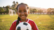 © Studio Nova - happy young girl with braided hair, holding a soccer ball, wearing a red sports jersey, with a soccer goal in the background, likely on a playing field