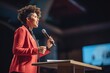 © lenblr - African-American woman leader gives keynote speech to audience at conference. Smiling woman speaker talks about business processes in microphone from podium