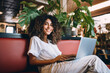 © Alfonso Soler - A joyful young latin american woman with curly hair comfortably uses a laptop.