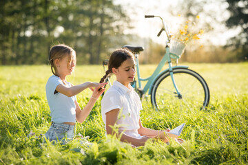 Wall Mural - Two teenage girls spend time on green grass lawn in park, braid pigtails and tails for each other, read books, enjoy summer and vacations