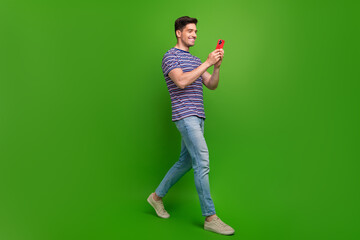 Poster - Full length photo of pleasant man wear t-shirt denim pants read email on smartphone walk empty space isolated on green color background