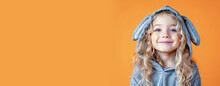 A Pretty Blonde Little Girl With A Sweet Smile In A Gray Easter Bunny Costume With Fluffy Ears On An Isolated Orange Background. Banner, A Place To Copy