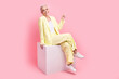 Full body photo of good mood person dressed yellow jacket indicating at promo empty space sit on podium isolated on pink color background